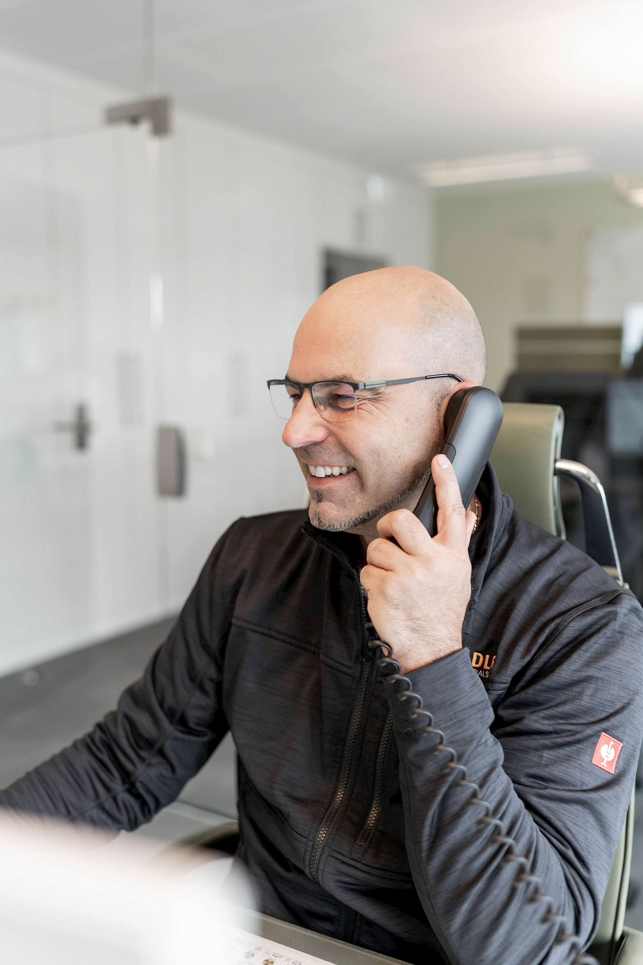 Ein Mann mit Brille sitzt in einem Büro und spricht lächelnd in ein Telefon.