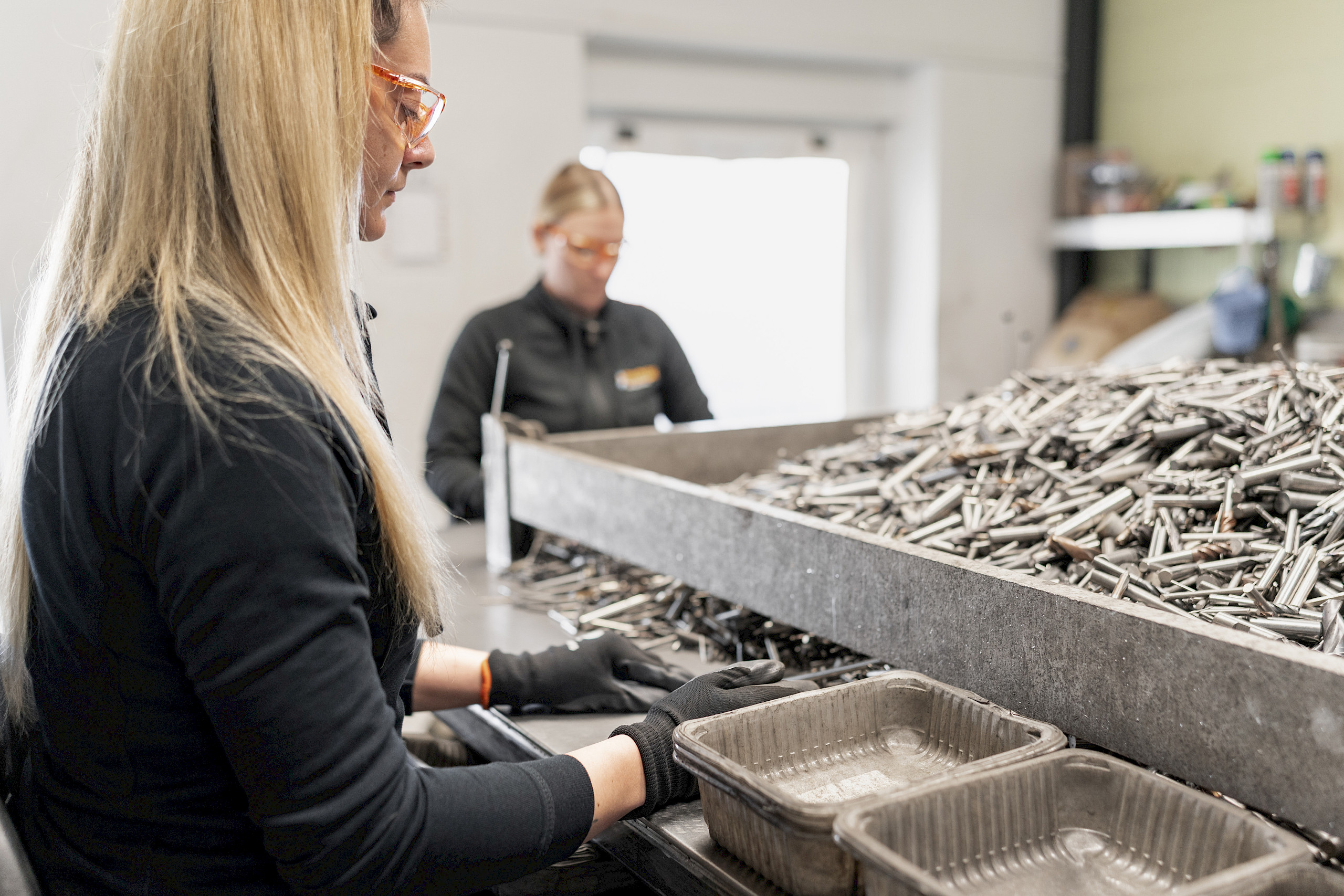 Frau sortiert Metallteile; im Hintergrund weitere Person; Innenaufnahme einer Werkstatt.