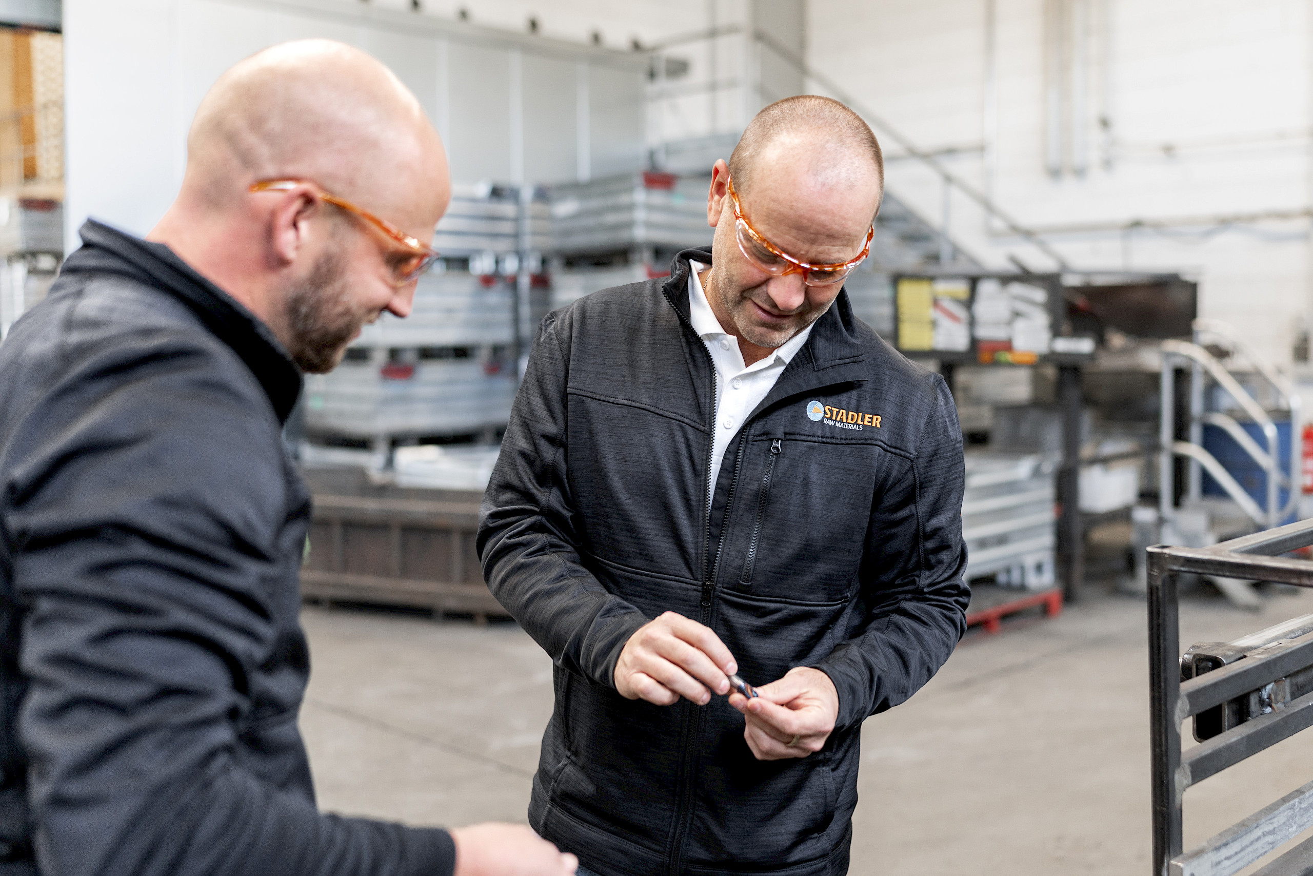 Zwei Männer in Arbeitskleidung und Schutzbrillen stehen in einer Werkstatt und betrachten gemeinsam einen kleinen Gegenstand.